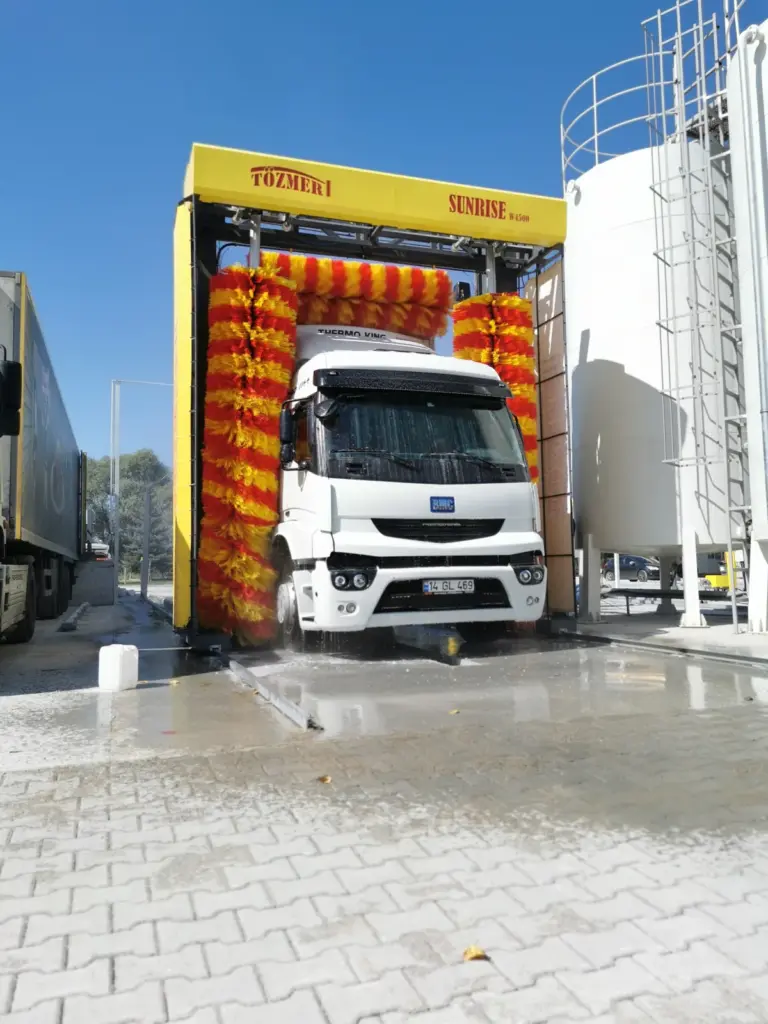 Red and yellow brushes closing on the cab of a white truck in an outdoor gantry, a silo to the right
